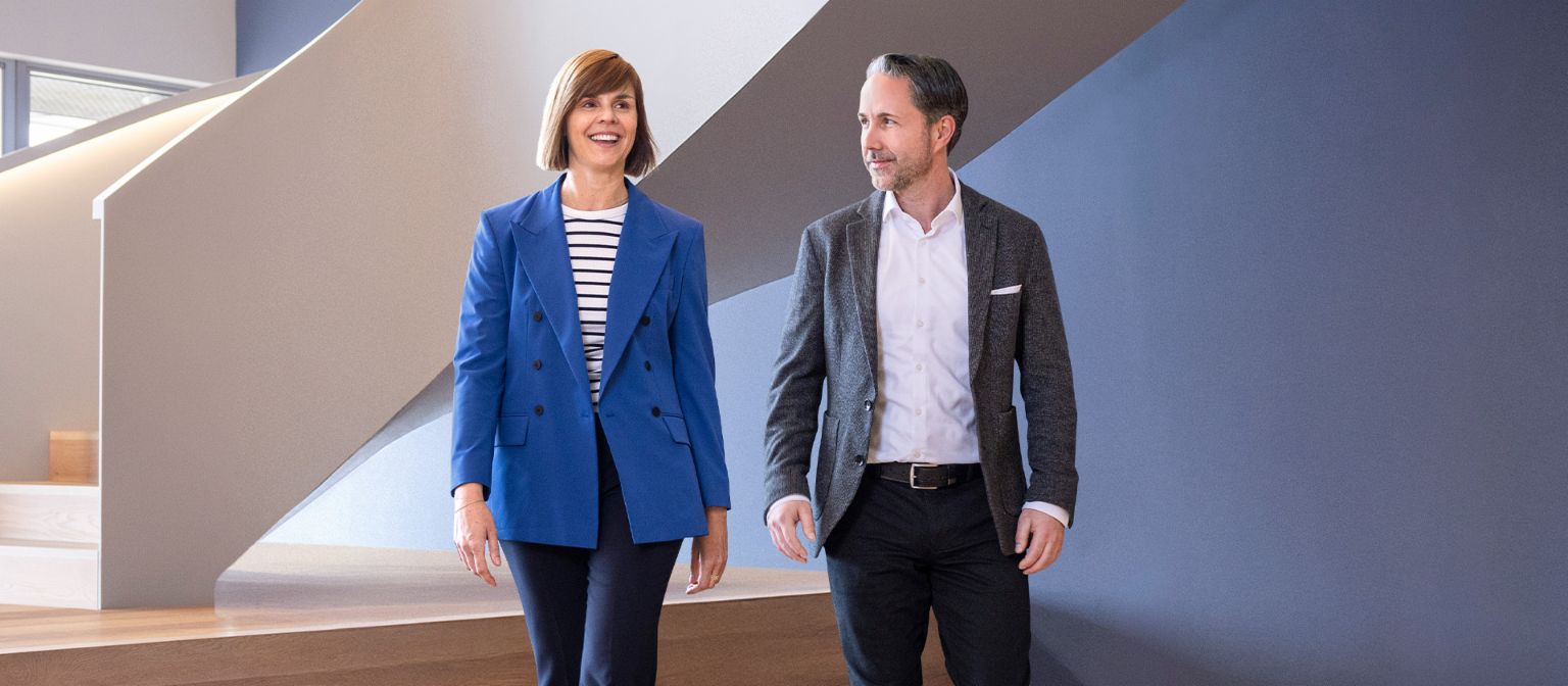 The CEO in a gray blazer and the CFO of the EOS Group in a blue blazer walk through a modern office. The space features large windows, a curved wall, and stylish hanging lamps.