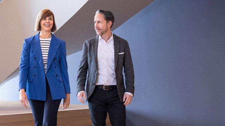 Der CEO im grauen Blazer und die CFO der EOS Gruppe in blauem Blazer gehen in einem modernen Büro. Der Raum hat große Fenster, eine geschwungene Wand und stilvolle hängende Lampen.