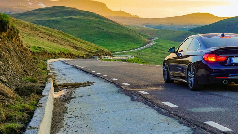 Eine Fotomontage zeigt ein Auto, das auf der rechten Spur einer asphaltierten Straße durch eine grüne, weite Landschaft in Richtung Sonnenuntergang fährt. Auf der entgegenkommenden Spur ist der Asphalt vollständig abgetragen.