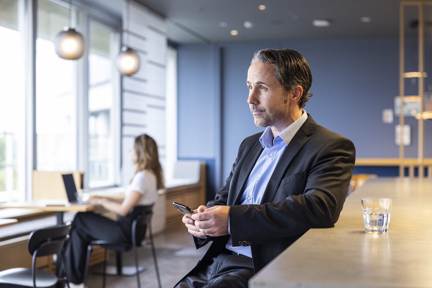 Marwin Ramcke is standing at the counter in a modern social space, wearing a black jacket, holding a glass of water and his phone.