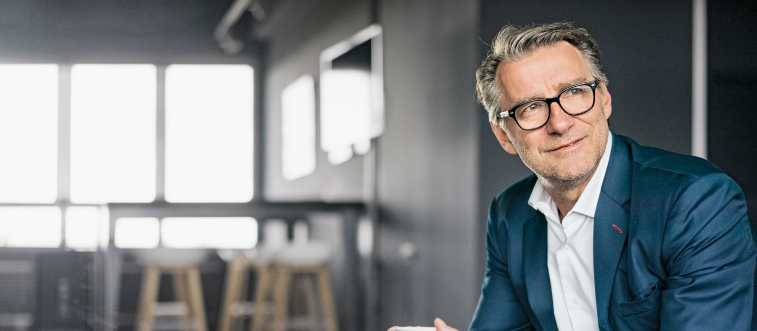  Portret zrelog poslovnog čovjeka koji sjedi, nosi plavo odijelo i drži bijelu šoljicu kafe.