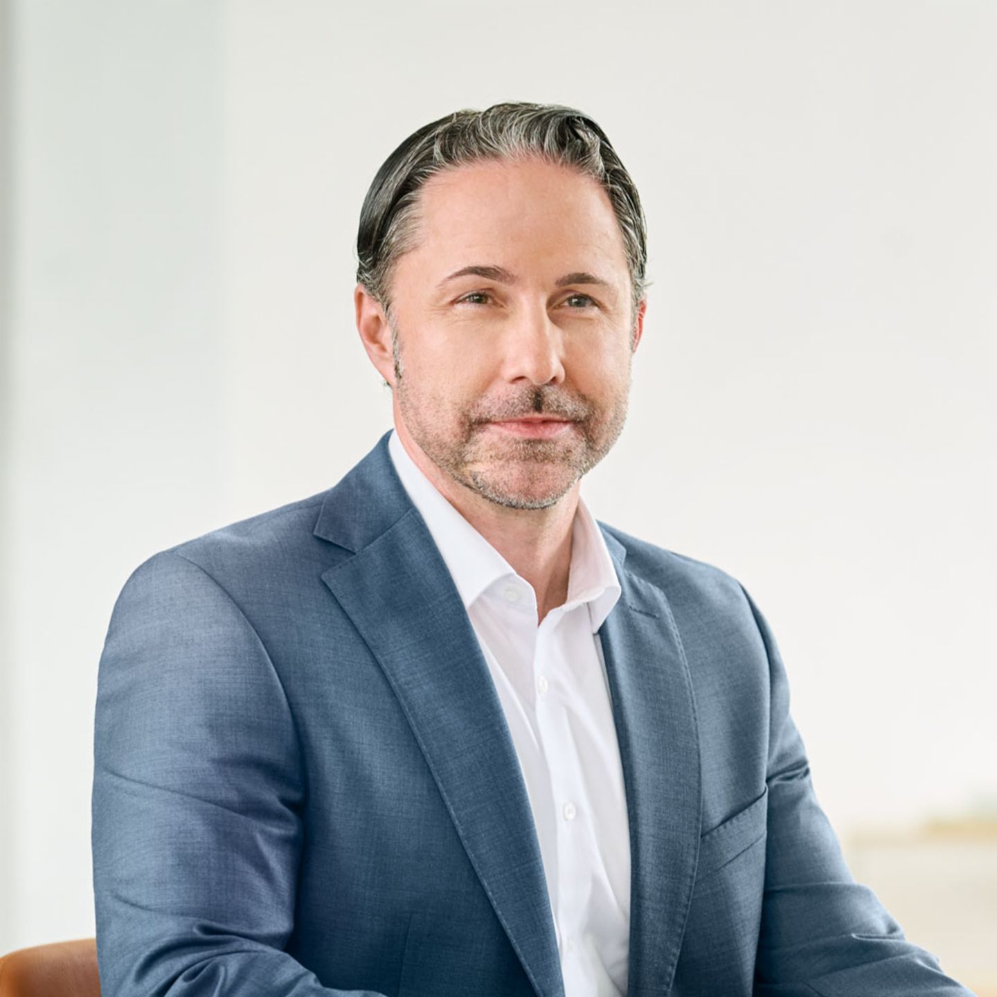 Marwin Ramcke, man in a dark blue suit and white shirt sitting on a chair in a bright room. 