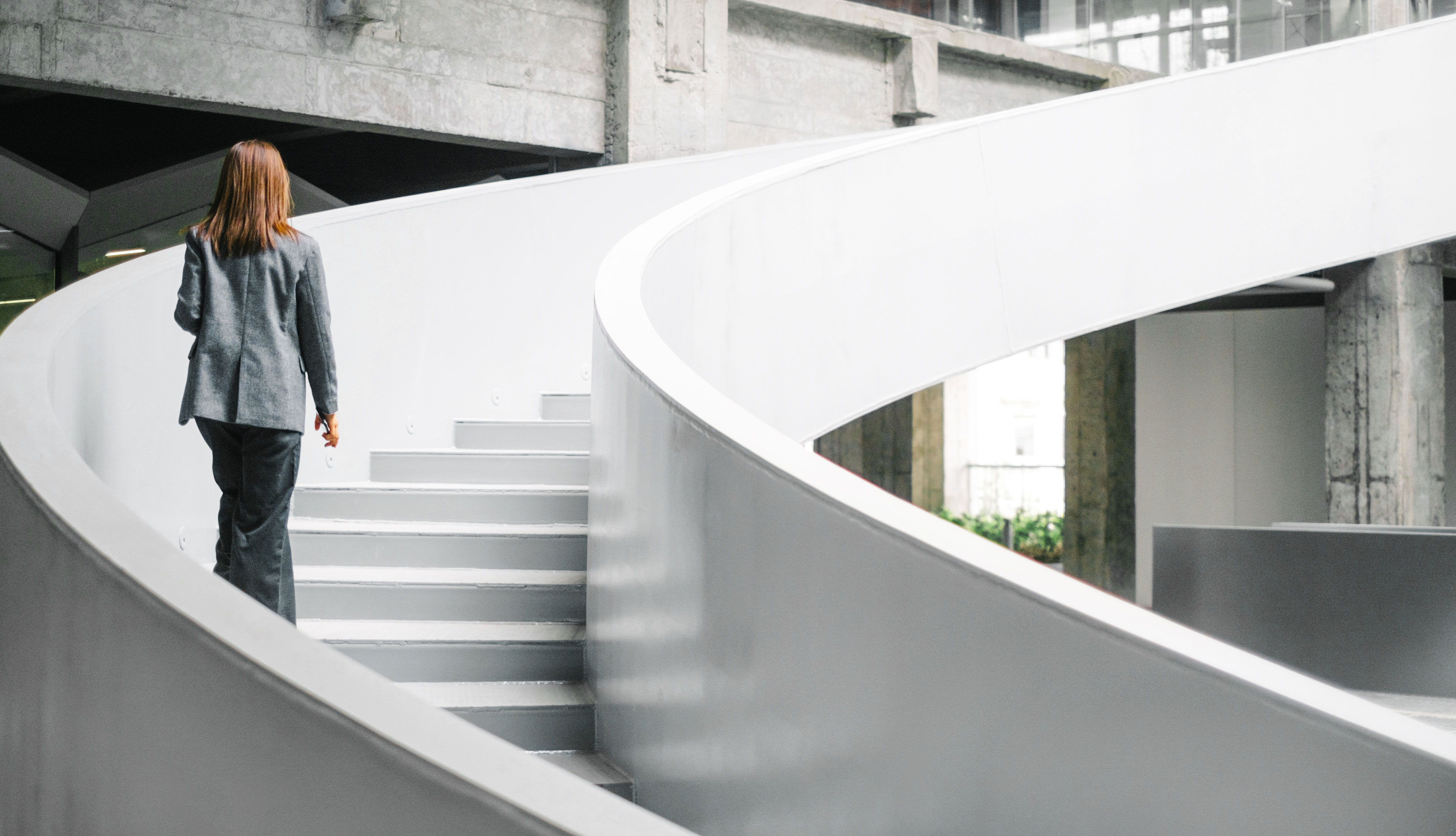 Businesswoman in a grey suit walking up white roud staircases the next flor in an office building