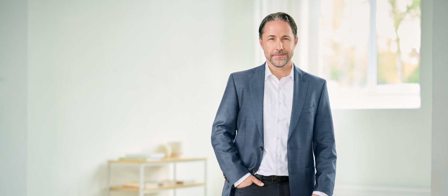 Marwin Ramcke, CEO, standing in a blue suit, with his right hand in his trouser pocket, in a bright room. 